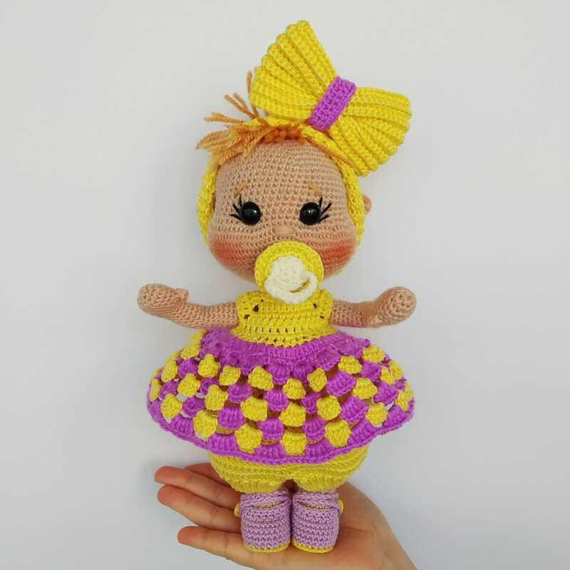 Amigurumi crochet baby doll with a large yellow bow, pacifier, and purple-and-yellow dress, shown in a hand for scale.