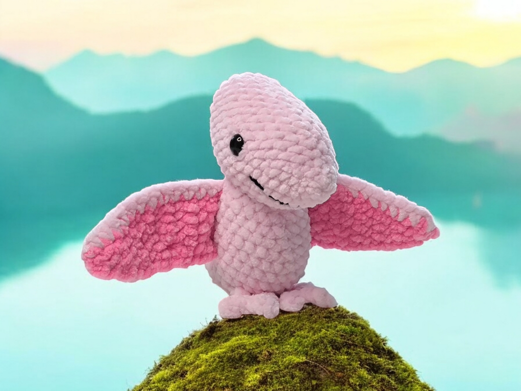 Rosa gehäkelter Flugsaurier mit hell- und dunkelrosa Flügeln auf einem Mooshügel, Berge und See im Hintergrund.
