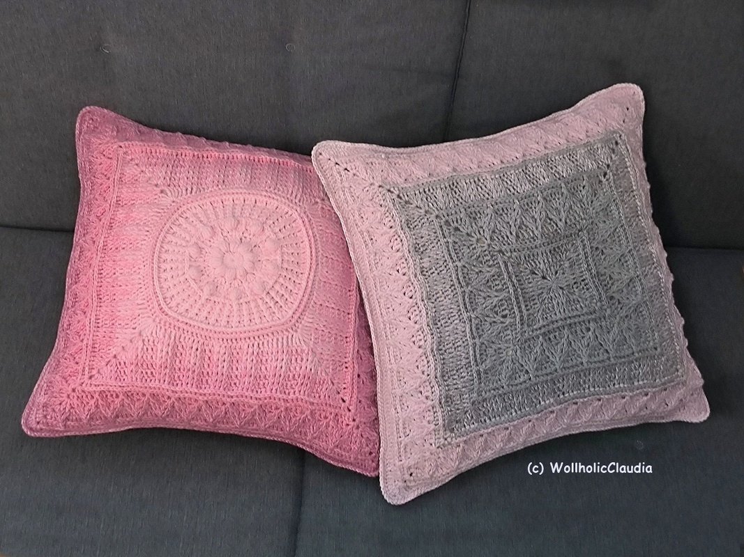 Zwei gehäkelte quadratische Kissenbezüge in Rosa und Grau mit dekorativer Zierkante auf einem dunklen Sofa