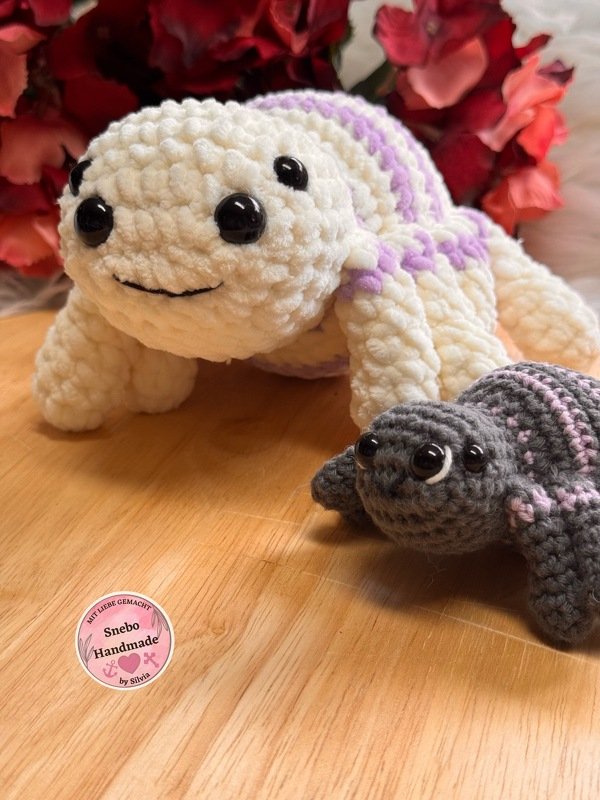 Zwei gehäkelte Amigurumi-Schildkröten (creme-lila und grau-rosa) auf einer Holzplatte vor roten Blumen.
