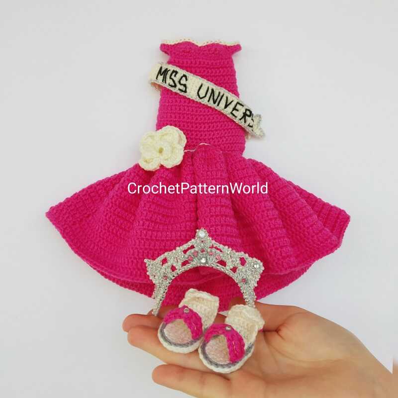 Bright pink crocheted doll dress with a cream sash and flower, a small decorative tiara, and two tiny pink-and-cream crocheted sandals held in a hand.