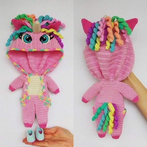 Front and back views of a pink crocheted unicorn doll jumpsuit with hood, colorful curly mane, small horn, tail, and a pair of light-blue shoes, held by a hand.