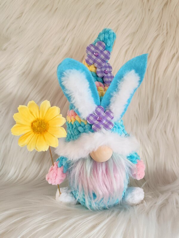 Gehäkelter Pastell-Wichtel mit Hasenohren, flauschigem Bart, gelber Blume und Mütze mit Stoffblüten vor hellem Hintergrund