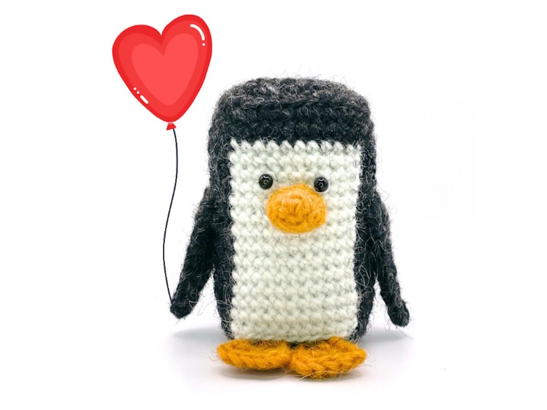 Gehäkelter Amigurumi-Pinguin in Schwarz, Weiß und Orange mit rotem Herzballon vor weißem Hintergrund.