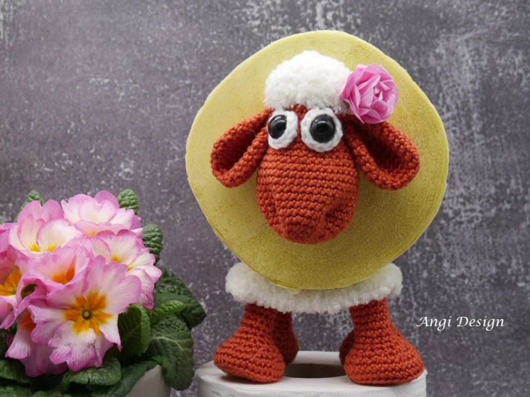 Gehäkelter Schaf-Toilettenpapierhalter mit orange-braunem Kopf, weißem Flausch und rosa Blüte neben pinken Primeln.