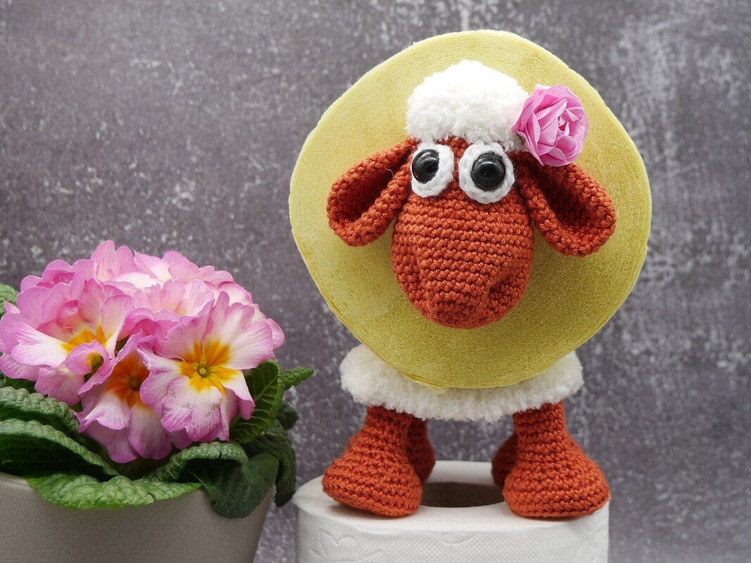 Gehäkeltes Schaf mit großen Augen, rosa Blume und gelbem Kreis, steht auf einer Toilettenpapierrolle neben einer Topfblume