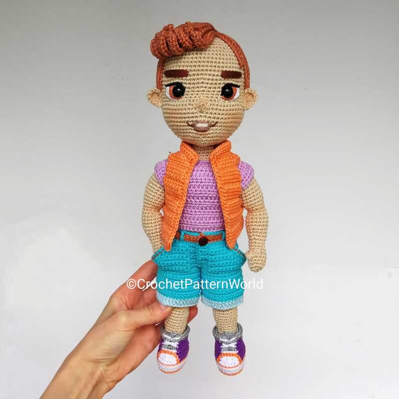 Crocheted amigurumi boy doll wearing orange vest, purple top, teal shorts, and two-tone sneakers, held by a hand