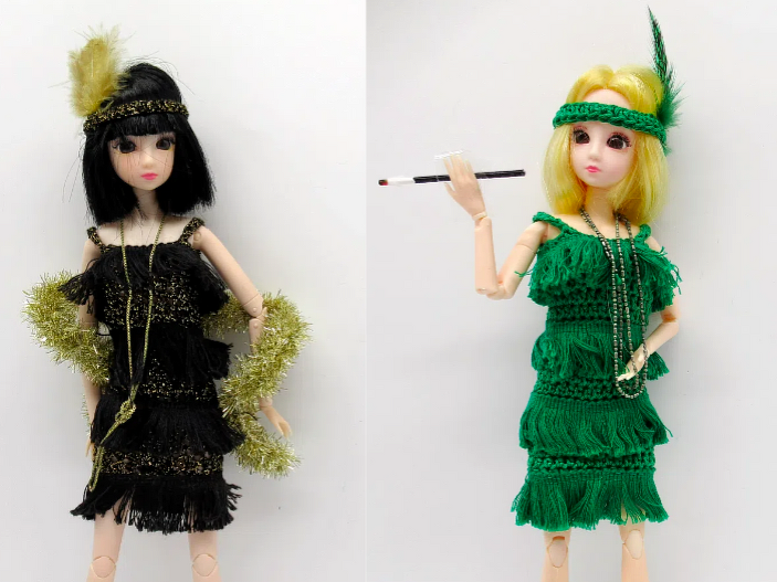 Zwei Puppen in gehäkelten Flapper‑Kleidern: links schwarz mit Goldfransen, rechts grün mit Federstirnband; grüne Puppe hält Zigarettenhalter.