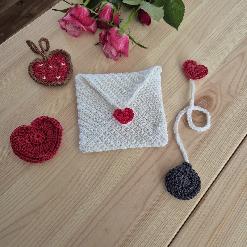 Verschiedene schnell gehäkelte Herzen, ein weißer Umschlag mit rotem Herzverschluss und ein Lesezeichen auf Holz