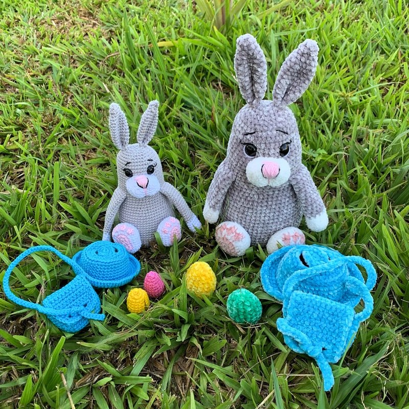 Two grey crocheted amigurumi rabbits on grass with small crocheted eggs and blue accessories