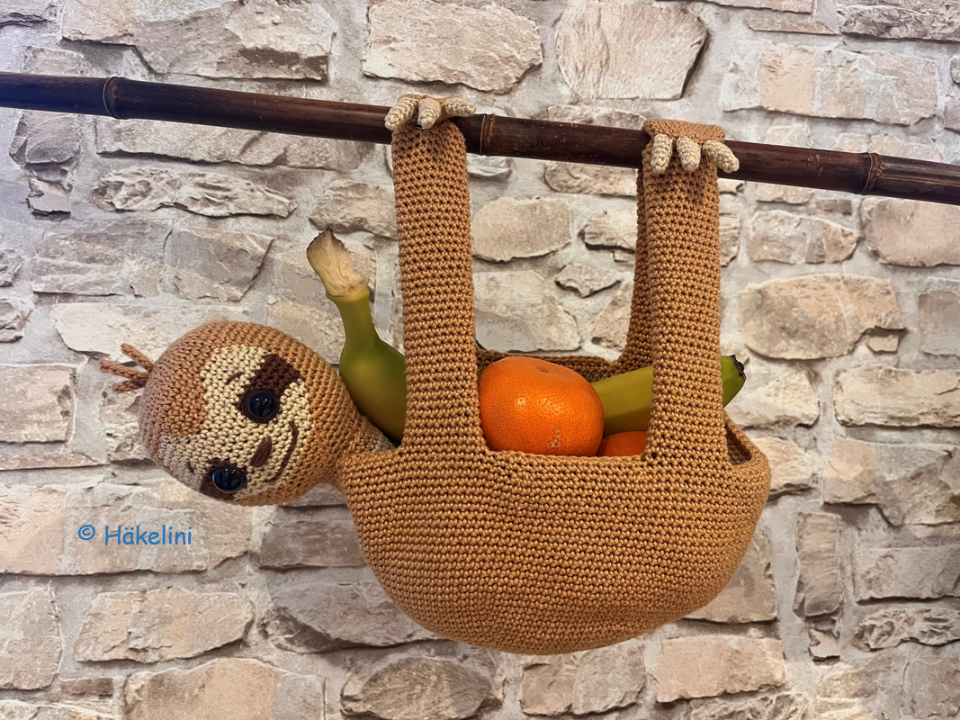 Brauner gehäkelter Faultier-Hängkorb hängt an Stange, gefüllt mit Bananen und Mandarinen