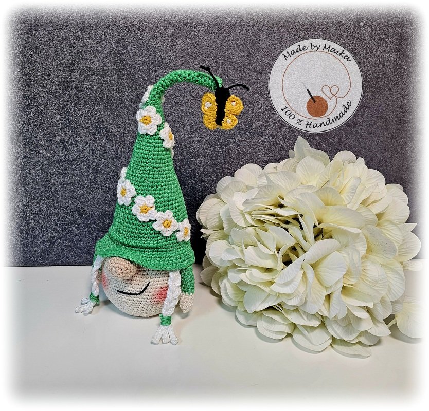 Amigurumi-Wichtel mit grünem Zipfelhut, weißen Gänseblümchen und kleinem gelben Schmetterling, neben einer großen weißen Blüte.