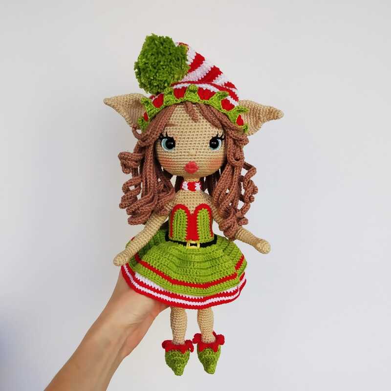 Hand holding a crocheted amigurumi Christmas elf doll with curly brown hair, a green dress, striped hat, and curled-toe shoes.