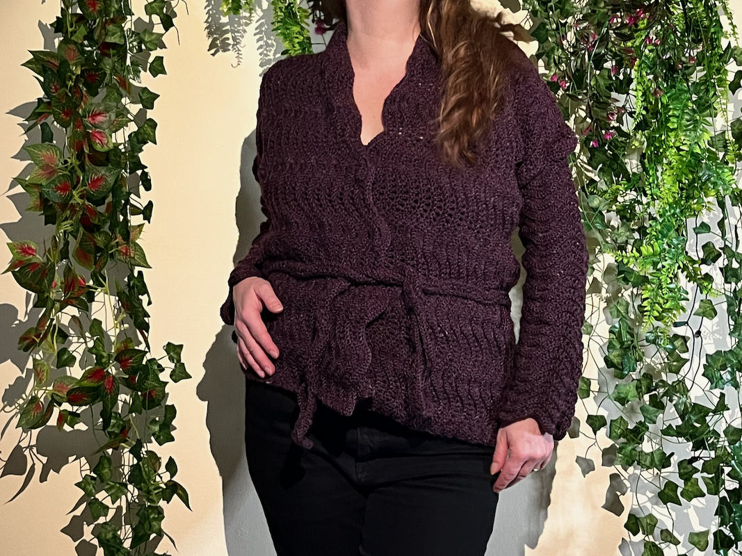Person wearing a dark plum crocheted jacket with tie belt, long sleeves and notched neckline against a leafy background