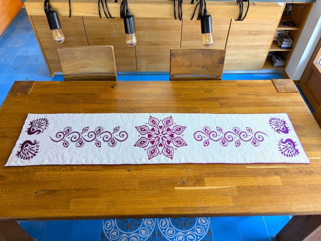 White knitted table runner with purple central mandala, scrollwork, and peacock motifs on a wooden dining table.