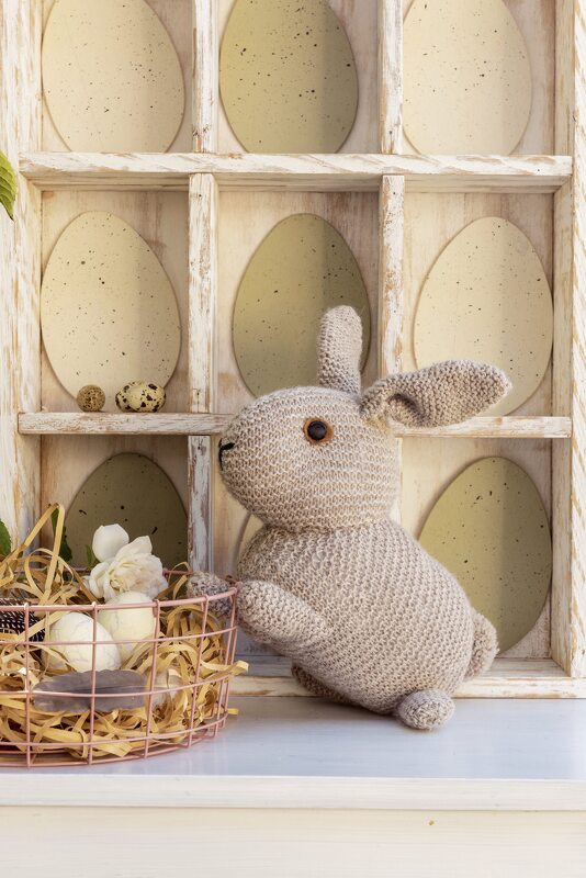 Grau gestrickter Amigurumi-Hase neben Drahtkorb mit Stroh, Eiern und Blumen vor weißgewaschenem Regal