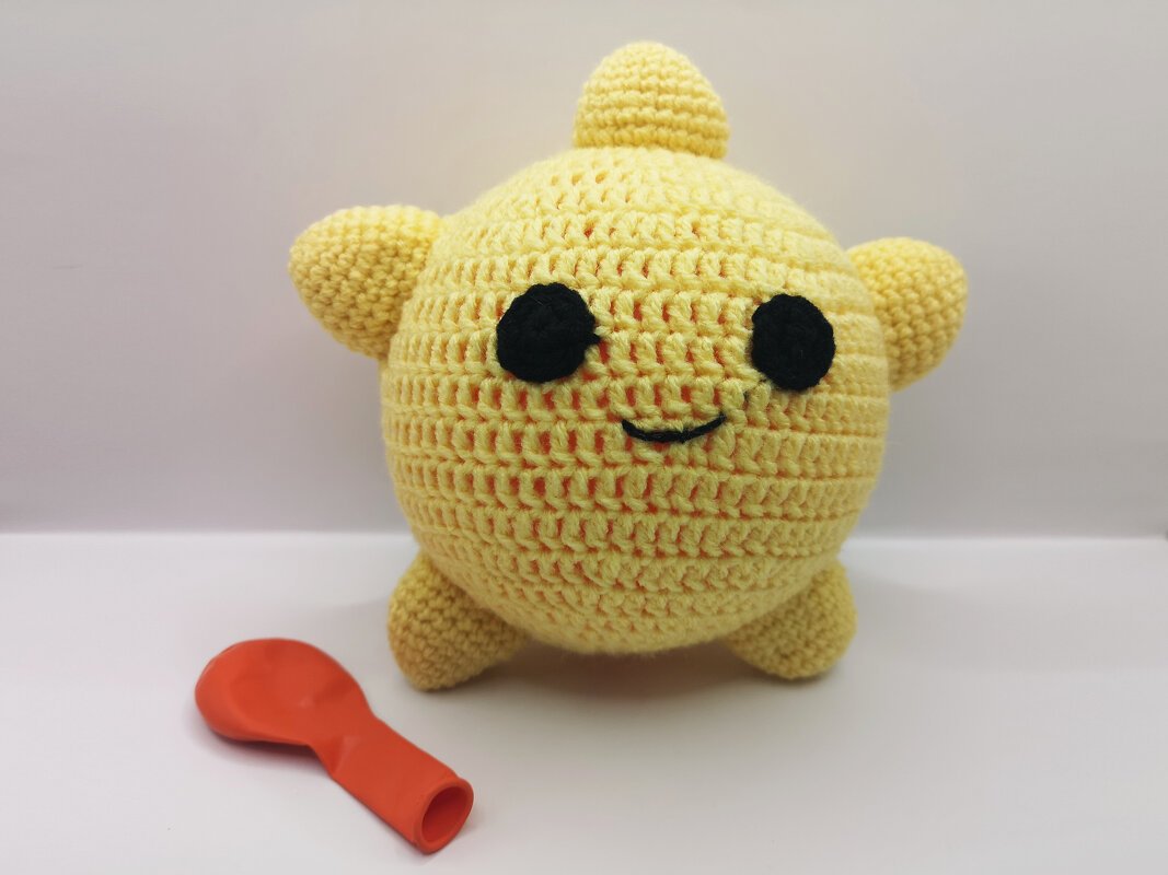 Gelbes gehäkeltes Amigurumi-Kuscheltier mit großen schwarzen Augen und orangefarbenem, unaufgeblasenen Ballon.