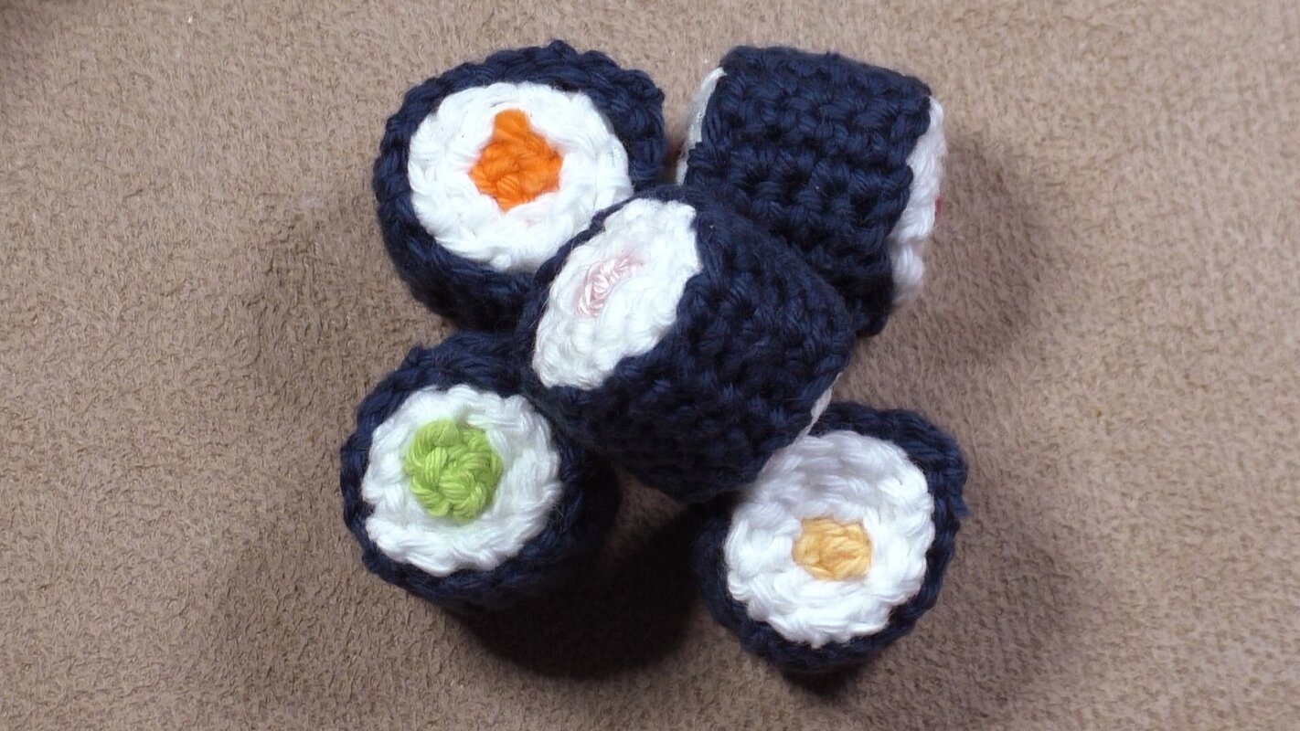 Fünf gehäkelte Sushi-Rollen mit dunkelblauer Umrandung, weißer Mitte und bunten Füllungen auf beigem Stoff.