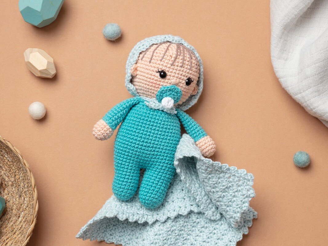 Gehäkelte Amigurumi-Babypuppe in türkis mit Schnuller und Häkeltuch auf beigem Hintergrund