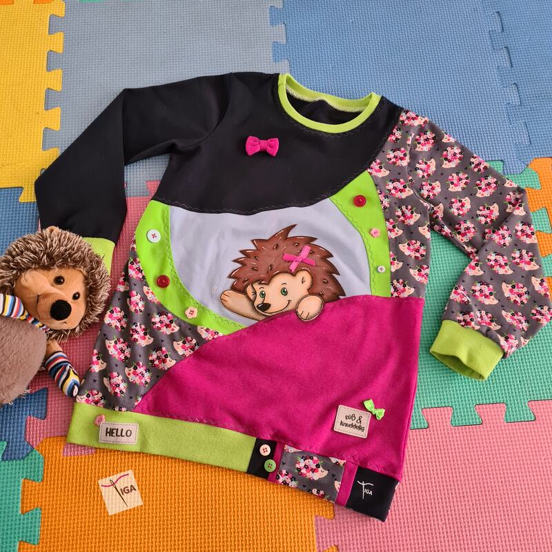 Asymmetrisches Kinder-Shirt liegend mit zentraler Igel-Applikation, pinkem Einsatz, grauem Ärmel mit Muster, grünen Bündchen und bunten Knöpfen; Plüsch-Igel daneben.