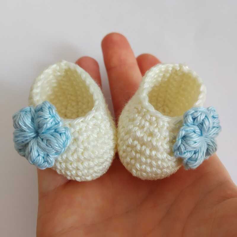 Winzige gehäkelte Babyschühchen in Creme mit hellblauer Blume auf einer Handfläche