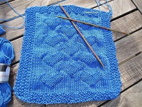 Blaues Strickquadrat mit eingestrickten Herzen, kraus-rechter Rand, Stricknadeln und Wollknäuel auf Holztisch