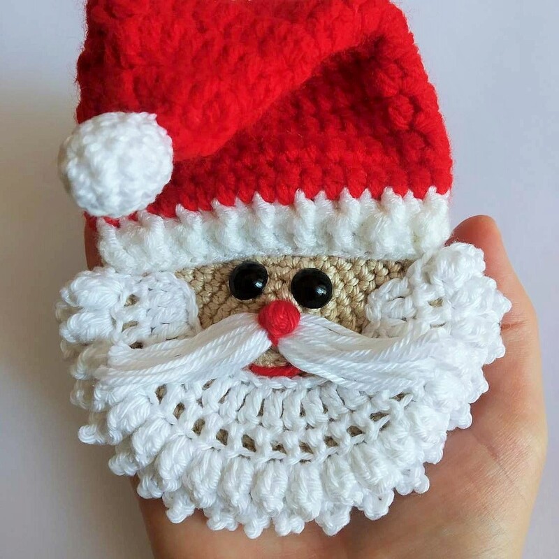 Small crocheted Santa Claus head with red hat, white beard and mustache, red nose and black round eyes held in a hand.