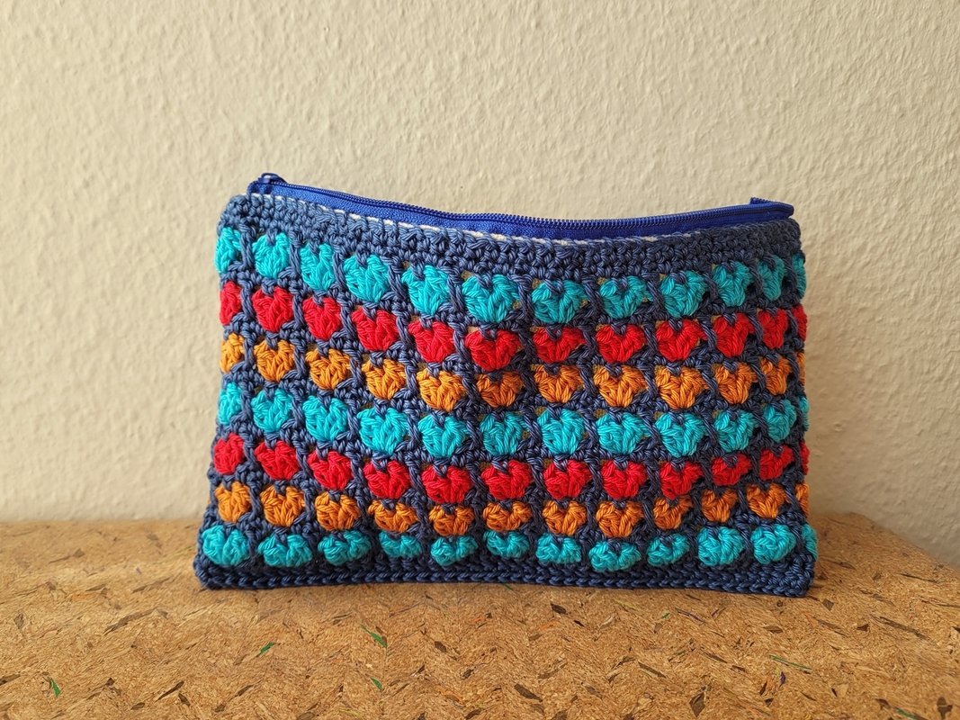 Gehäkeltes Täschchen mit Reihen kleiner Herzen in Türkis, Rot und Orange auf dunkelblauem Grund und blauem Reißverschluss