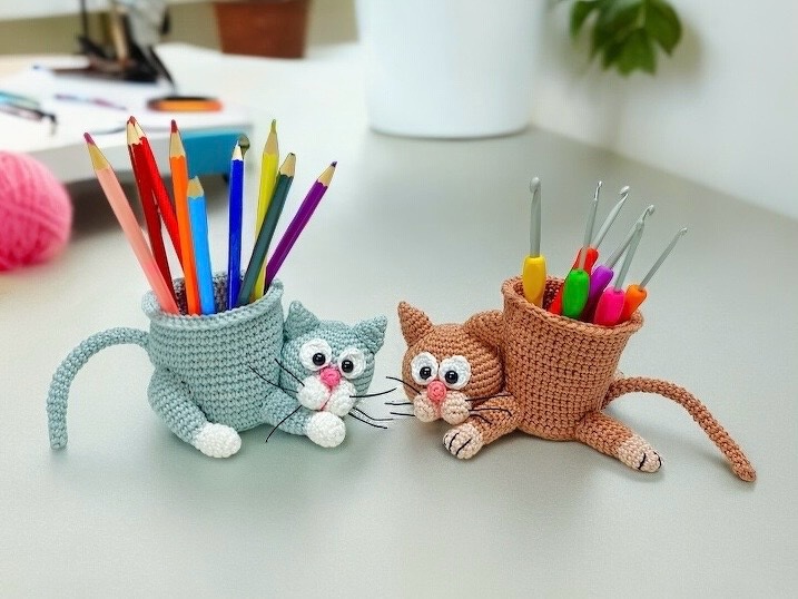 Zwei gehäkelte Katzen-Utensilos (hellblau und braun) mit Buntstiften und Häkelnadeln auf einem Schreibtisch.