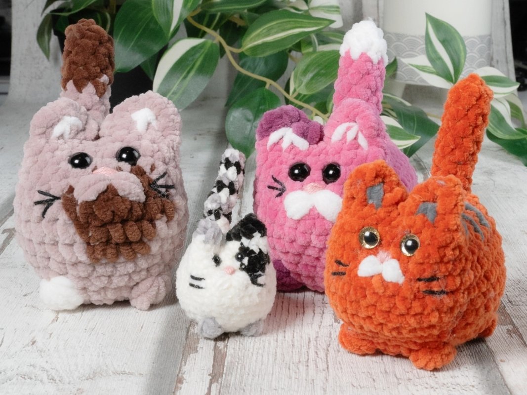 Vier flauschige gehäkelte Amigurumi-Katzen in Rosé, Pink, Orange und Weiß-Schwarz auf hellem Holztisch.