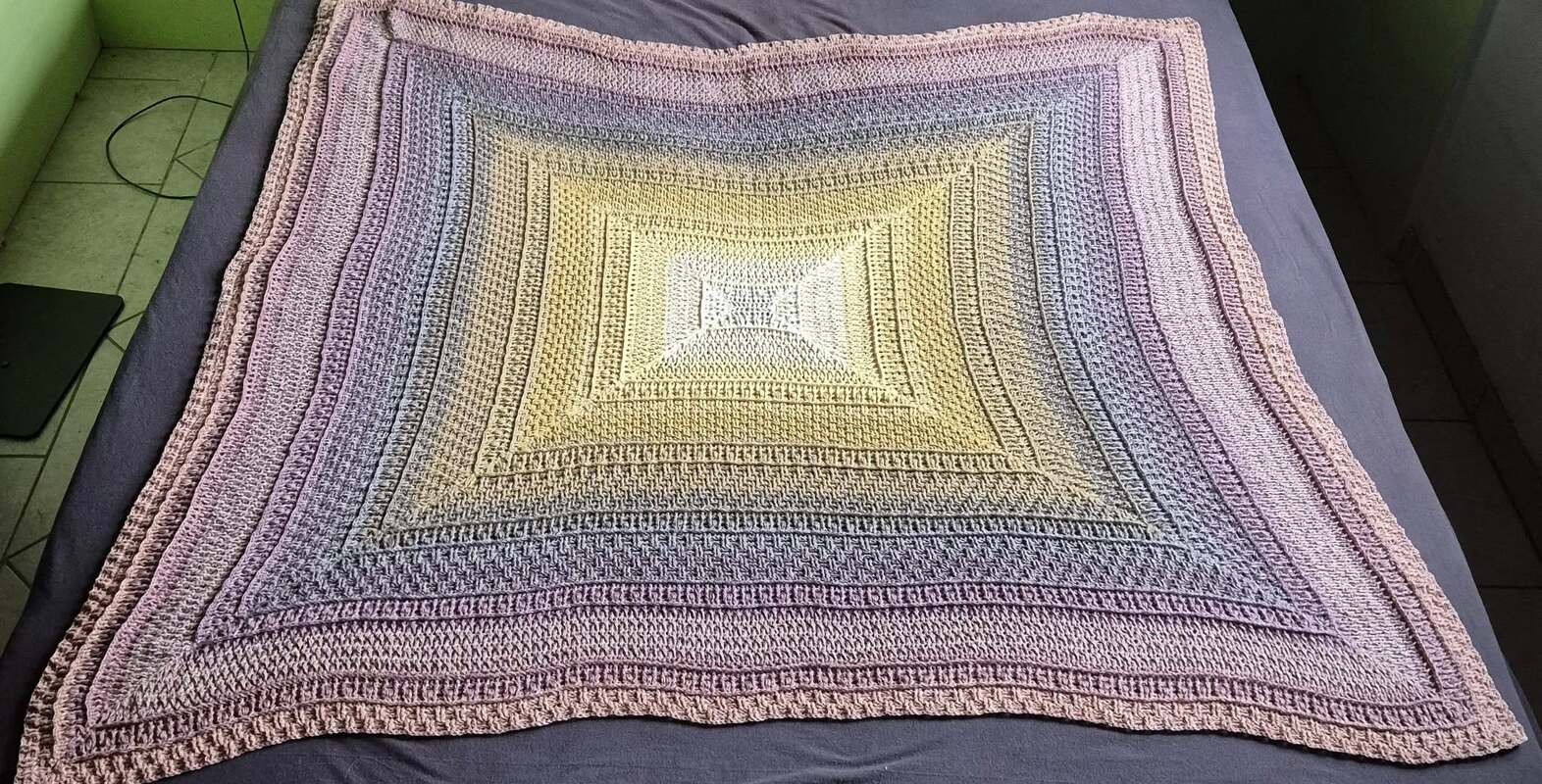 Quadratische Häkeldecke mit sanftem Verlauf von Hellgelb zu Blau-, Lila- und Rosatönen auf dunkelgrauem Untergrund.