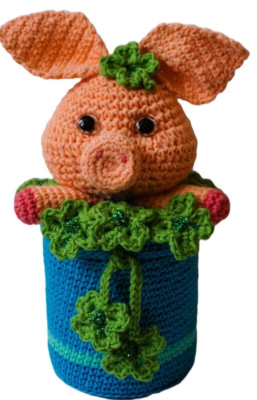 Gehäkeltes oranges Amigurumi-Schwein mit schwarzen Sicherheitsaugen in blauem Topf, grüne gehäkelte Kleeblätter als Verzierung