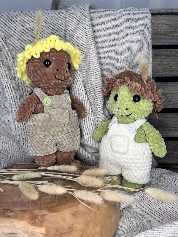 Zwei gehäkelte Amigurumi aus Plüschgarn mit Latzhosen und Knöpfen vor Stoff drapiert