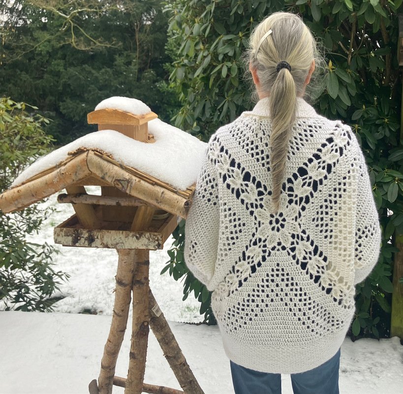 Rückansicht einer Person mit cremefarbenem gehäkeltem Seelenwärmer im verschneiten Garten neben einem Vogelfutterhaus.