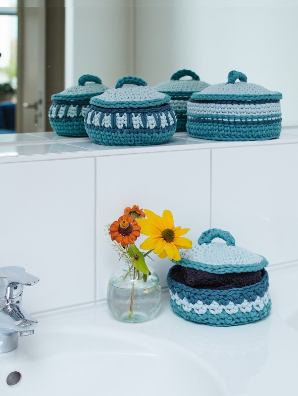 Gehäkelte Körbe mit Deckel in Blau- und Türkistönen, gefüllt mit Handtüchern neben einer Vase auf der Badablage
