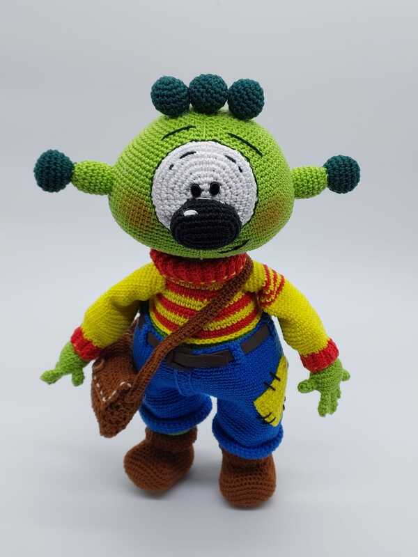 Gehäkeltes grünes Amigurumi-Monster mit großer Augenpartie, Fühlern, gestreiftem Shirt, blauer Hose, Stiefeln und Umhängetasche