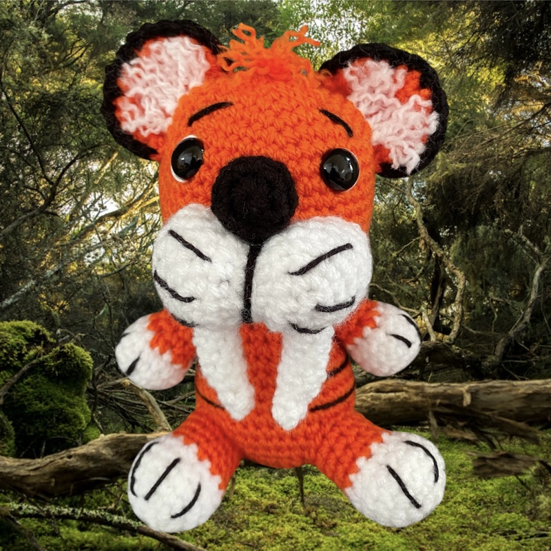Gehäkeltes orange-weißes Amigurumi mit Stoßzähnen und schwarzen Knopfaugen vor bewaldetem Hintergrund