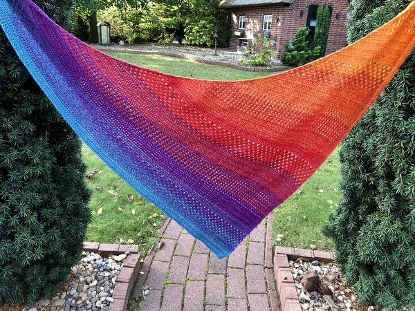 Gehäkeltes Dreieckstuch in leuchtenden Blau-, Violett- und Orangetönen mit filigranem Lochmuster, im Garten zwischen Sträuchern aufgehängt.