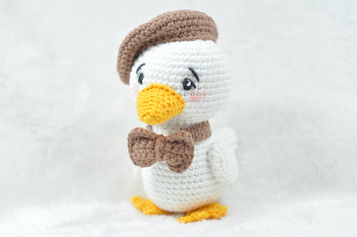 White crocheted amigurumi duck wearing a brown beret and bow tie with yellow beak and feet