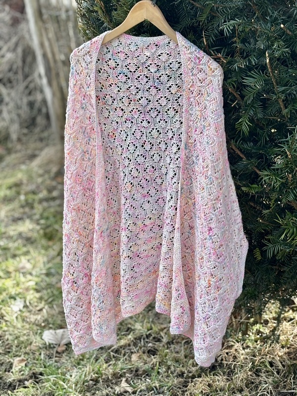 Offene, gestrickte Damenjacke in Rosa mit mehrfarbigem Ajourmuster, an einem Holzbügel vor grünen Büschen hängend.