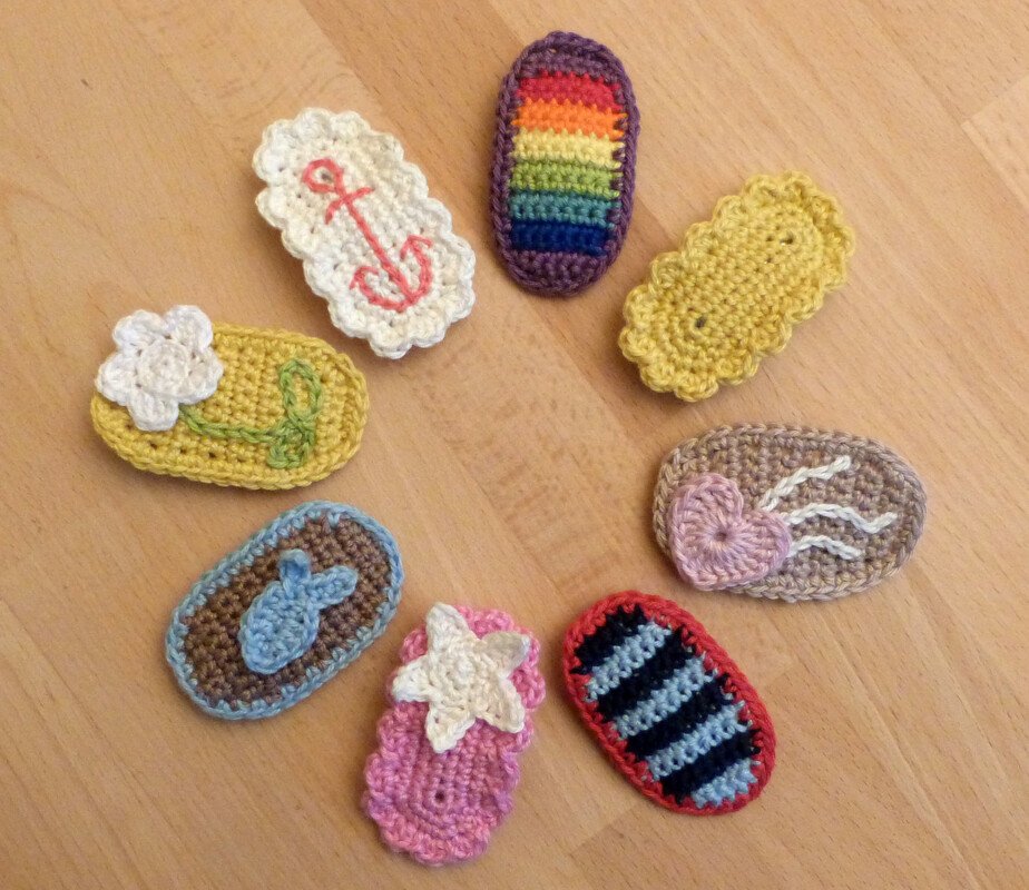 Neun kleine gehäkelte Applikationen auf Holzuntergrund mit Motiven wie Regenbogen, Anker, Blume, Herz, Stern und Streifen.