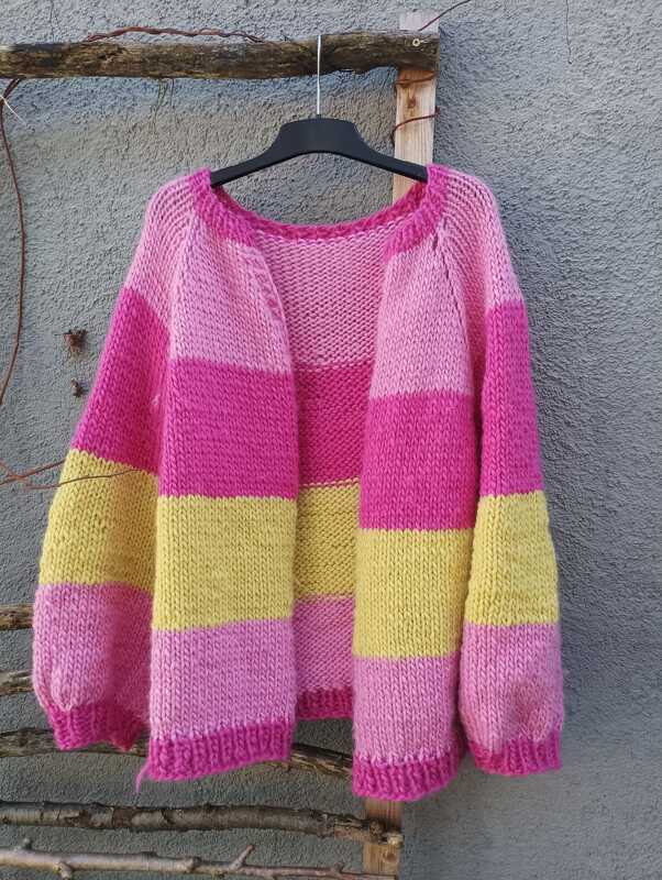 Grobgestrickte Damenjacke mit breiten pinken und gelben Streifen an einem schwarzen Bügel vor grauer Wand