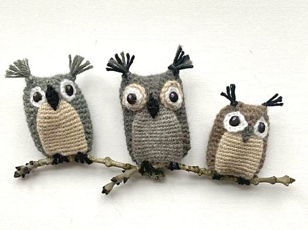 Drei gehäkelte Amigurumi-Eulen in Grau- und Brauntönen mit beigen Bäuchen sitzen auf einem Zweig.