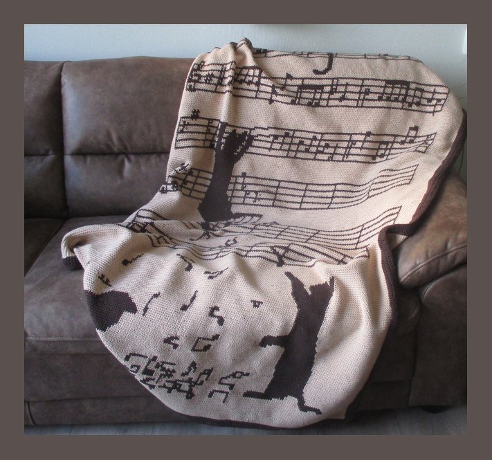 Beige gehäkelte Decke mit schwarzen Musiknoten und Katzen auf einem braunen Sofa