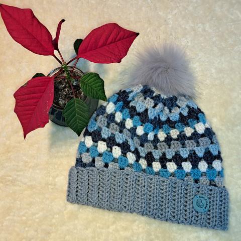 Gehäkelte Mütze mit Granny-Square-Muster in Blau-, Grau- und Weißtönen und grauem Bommel; ein Weihnachtsstern daneben.