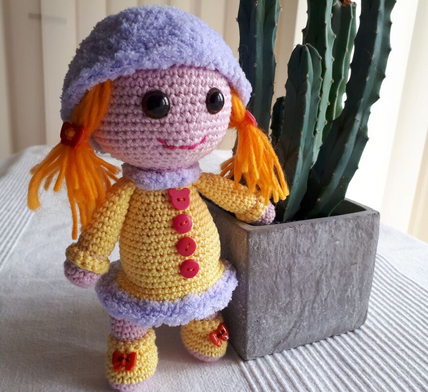 Gehäkelte Amigurumi-Puppe mit orangeroten Zöpfen, lila Mütze und gelbem Mantel neben einem Kaktus.