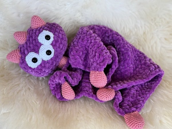 Violettes gehäkeltes Schmusetuch mit Monsterkopf, weißen Augen und rosa Knotenenden auf hellem, flauschigem Untergrund.