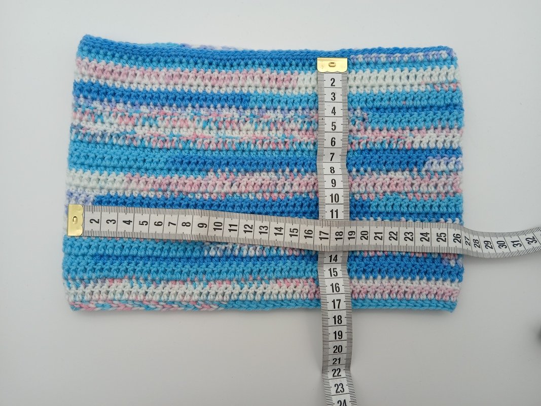 Gehäkelter Loop-Schal mit Streifen in Blau, Rosa und Weiß, zwei Maßbänder liegen über Kreuz zur Größenangabe.