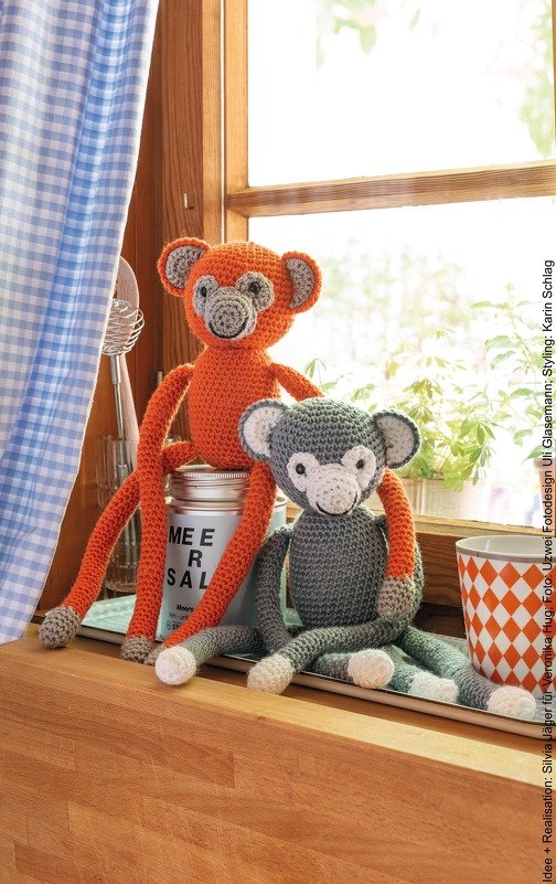 Zwei Amigurumi-Affen mit langen Armen und kontrastierenden Pfoten sitzen nebeneinander auf einer Fensterbank