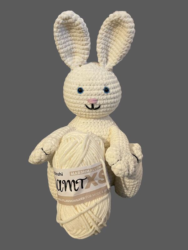 Weißer gehäkelter Amigurumi-Hase mit blauen Augen und rosa Nase, der ein cremefarbenes Garnknäuel hält.